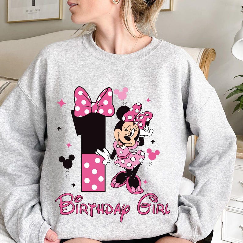 May include: A gray sweatshirt featuring a graphic of Minnie Mouse, a large number 1, and the text "Birthday Girl" in pink. Minnie Mouse is wearing a pink polka dot dress and bow. The number 1 has pink polka dots.