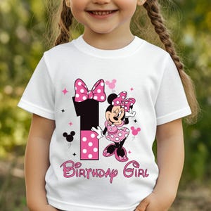May include: White t-shirt featuring a Minnie Mouse graphic with a pink polka dot bow and dress. The number 1 is in black with pink polka dots. The words "Birthday Girl" are in pink script.