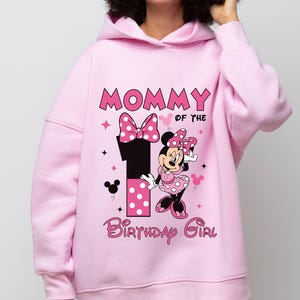 May include: Pink hooded sweatshirt with the text "MOMMY OF THE 1 BIRTHDAY GIRL" and a Minnie Mouse graphic. The graphic includes a large number 1, a bow, and Minnie Mouse in a polka dot dress. The design is in pink, black, and white.