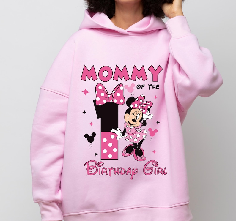 May include: Pink hooded sweatshirt with the text "MOMMY OF THE 1 BIRTHDAY GIRL" and a Minnie Mouse graphic. The graphic includes a large number 1, a bow, and Minnie Mouse in a polka dot dress. The design is in pink, black, and white.