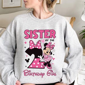 Puede incluir: Una sudadera gris claro con un gr&aacute;fico rosa y negro. El gr&aacute;fico presenta el texto "SISTER OF THE BIRTHDAY GIRL" y un dise&ntilde;o de Minnie Mouse con un lazo y un vestido de lunares rosas. El n&uacute;mero "2" tambi&eacute;n est&aacute; en el dise&ntilde;o.