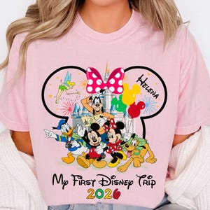 May include: Pink t-shirt featuring a Disney-themed graphic. The design includes Mickey Mouse, Minnie Mouse, Donald Duck, Goofy, and Pluto, with a castle backdrop, balloons, and the text "My First Disney Trip 2020".