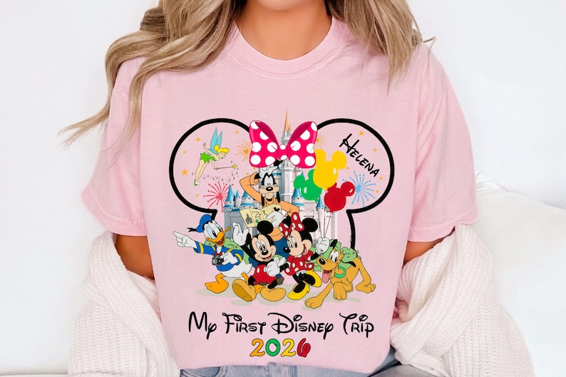 May include: Pink t-shirt featuring a Disney-themed graphic. The design includes Mickey Mouse, Minnie Mouse, Donald Duck, Goofy, and Pluto, with a castle backdrop, balloons, and the text "My First Disney Trip 2020".