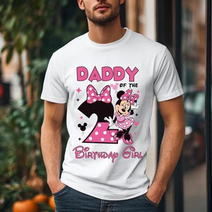 Puede incluir: Camiseta blanca con un gr&aacute;fico rosa que dice "DADDY OF THE BIRTHDAY GIRL" y presenta un dise&ntilde;o de Minnie Mouse. El n&uacute;mero "2" tambi&eacute;n est&aacute; en el dise&ntilde;o, con un patr&oacute;n de lunares. La camiseta es de manga corta.