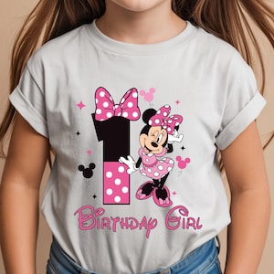 May include: A light gray t-shirt featuring a Minnie Mouse design for a first birthday. The design includes a large number 1, Minnie Mouse, pink polka dots, stars, and the words "Birthday Girl" in pink script.