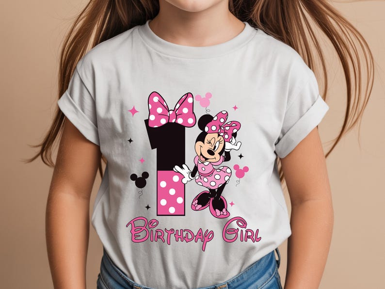 May include: A light gray t-shirt featuring a Minnie Mouse design for a first birthday. The design includes a large number 1, Minnie Mouse, pink polka dots, stars, and the words "Birthday Girl" in pink script.