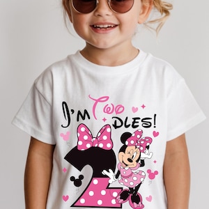 May include: White t-shirt with a Minnie Mouse graphic for a two-year-old. The design includes a large number 2 with pink polka dots, a pink bow, and the text "I'm Two Dles!" in black and pink. The child is wearing sunglasses.