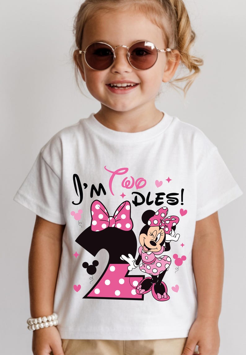 May include: White t-shirt with a Minnie Mouse graphic for a two-year-old. The design includes a large number 2 with pink polka dots, a pink bow, and the text "I'm Two Dles!" in black and pink. The child is wearing sunglasses.