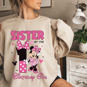 May include: A beige sweatshirt with a pink Minnie Mouse design. The design includes the words "SISTER OF THE" above a large number "1" and "BIRTHDAY GIRL" in a script font. Minnie Mouse is wearing a pink polka dot dress.