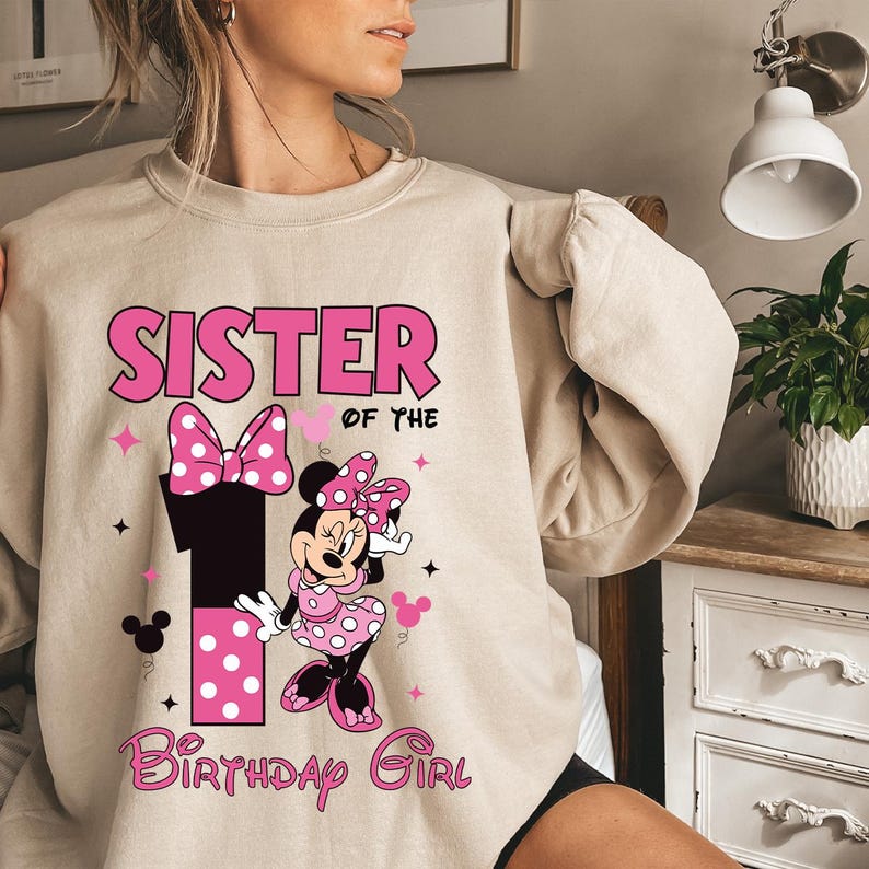 May include: A beige sweatshirt with a pink Minnie Mouse design. The design includes the words "SISTER OF THE" above a large number "1" and "BIRTHDAY GIRL" in a script font. Minnie Mouse is wearing a pink polka dot dress.