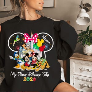 May include: Black sweatshirt featuring a colorful graphic of Disney characters including Mickey and Minnie Mouse, Donald Duck, and Goofy. The design includes the text "My First Disney Trip 2020" and the name "Helena". A pink polka dot bow is also featured.
