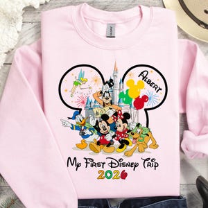May include: A light pink sweatshirt featuring a graphic of Disney characters, including Mickey and Minnie Mouse, Donald Duck, and Goofy. The design includes the text "My First Disney Trip 2020" and the name "Albert".