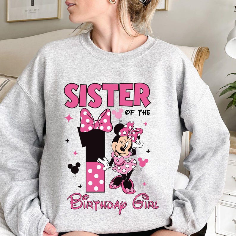 May include: A light gray sweatshirt with the text "SISTER OF THE BIRTHDAY GIRL" in pink. The design includes a large number 1 with pink polka dots, a Minnie Mouse graphic, and pink bows and stars.