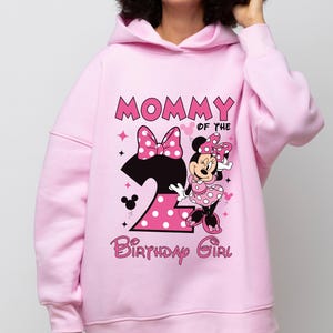 Puede incluir: Sudadera con capucha rosa claro con un dise&ntilde;o de Minnie Mouse y el texto "MOMMY OF THE BIRTHDAY GIRL". Tambi&eacute;n se muestra el n&uacute;mero "2", junto con un lazo y lunares. La sudadera est&aacute; hecha de un material suave.