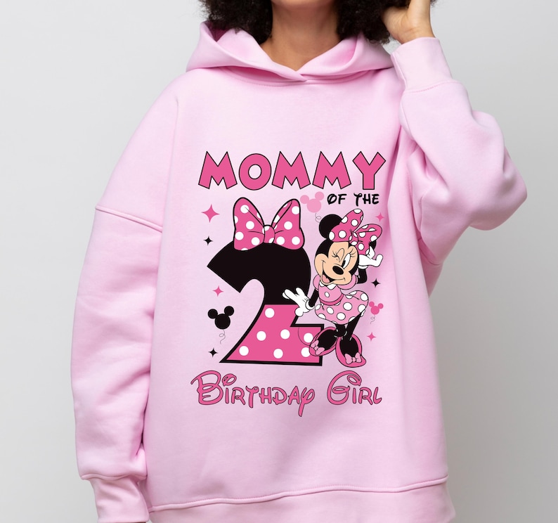 Puede incluir: Sudadera con capucha rosa claro con un dise&ntilde;o de Minnie Mouse y el texto "MOMMY OF THE BIRTHDAY GIRL". Tambi&eacute;n se muestra el n&uacute;mero "2", junto con un lazo y lunares. La sudadera est&aacute; hecha de un material suave.
