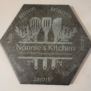 May include: A hexagonal slate tile with the words "Nonnie's Kitchen" and "where the only thing spoiled are the grandkids" engraved in white. The tile is decorated with a white silhouette of kitchen utensils and floral sprigs. The names Allison, Brianna, Hazel, Henry, Jackson, Leo, and Violet are engraved around the perimeter of the tile.