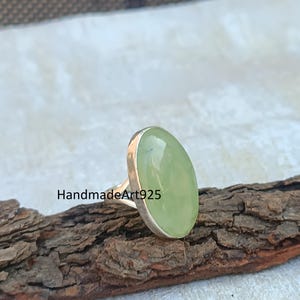 K&ouml;nnte beinhalten: Ein silberner Ring mit einem gro&szlig;en, ovalen, gr&uuml;nen Edelstein. Der Ring liegt auf einem St&uuml;ck Holz.