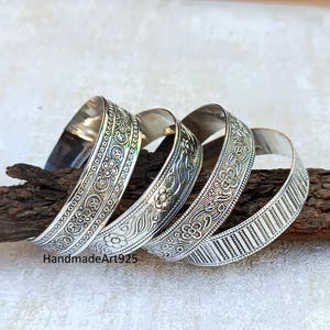 Może przedstawiać: Cztery srebrne bransoletki z misternymi wzorami kwiatowymi i geometrycznymi. Bransoletki są ułożone jedna na drugiej i mają błyszczące wykończenie.