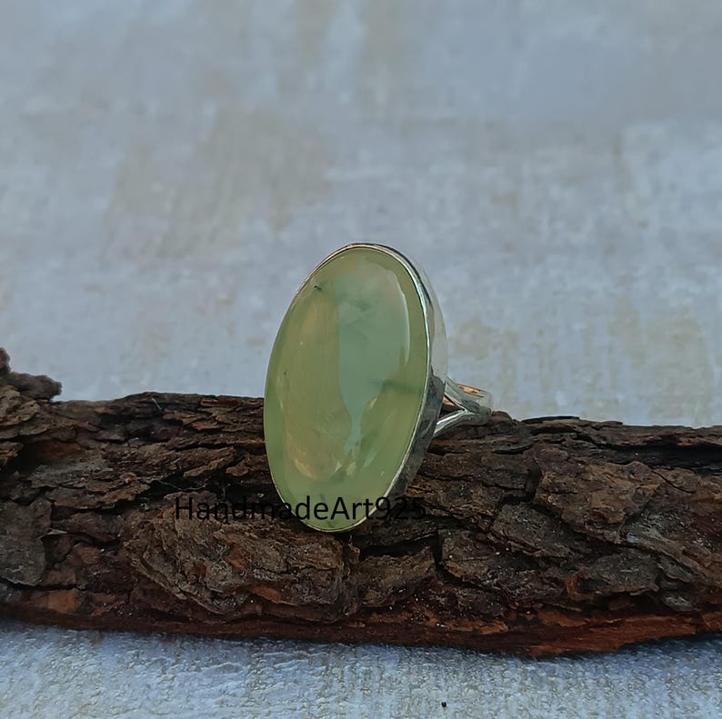 K&ouml;nnte beinhalten: Ein silberner Ring mit einem gro&szlig;en ovalen gr&uuml;nen Edelstein. Der Ring liegt auf einem St&uuml;ck Holz.