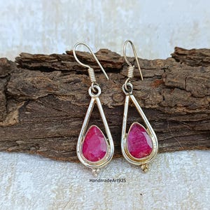 May include: A pair of silver teardrop earrings with vibrant red gemstones. The earrings feature a polished silver frame surrounding the faceted ruby-colored stones. The text "HandmadeArt925" is visible on the bottom.