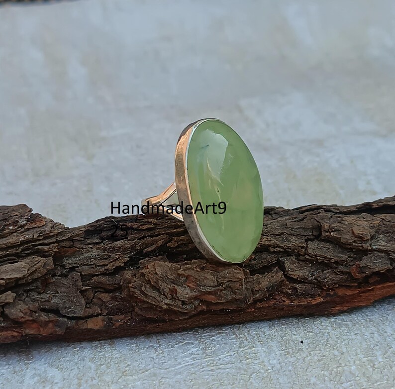 K&ouml;nnte beinhalten: Ein silberner Ring mit einem hellgr&uuml;nen ovalen Edelstein. Der Ring liegt auf einem St&uuml;ck Holz.
