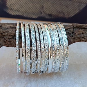 Peut inclure: Un ensemble de 12 bracelets en argent avec différents motifs en relief. Les bracelets sont empilés sur une surface en bois.
