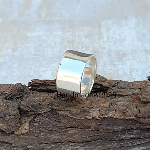 May include: A silver band ring with a wide, flat, polished surface. The ring is sitting on a piece of wood.