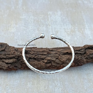 Peut inclure: Un bracelet jonc en argent martelé avec des extrémités arrondies. Le bracelet est posé sur une surface en bois.