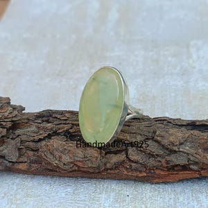 K&ouml;nnte beinhalten: Ein silberner Ring mit einem gro&szlig;en ovalen gr&uuml;nen Edelstein. Der Ring liegt auf einem St&uuml;ck Holz.