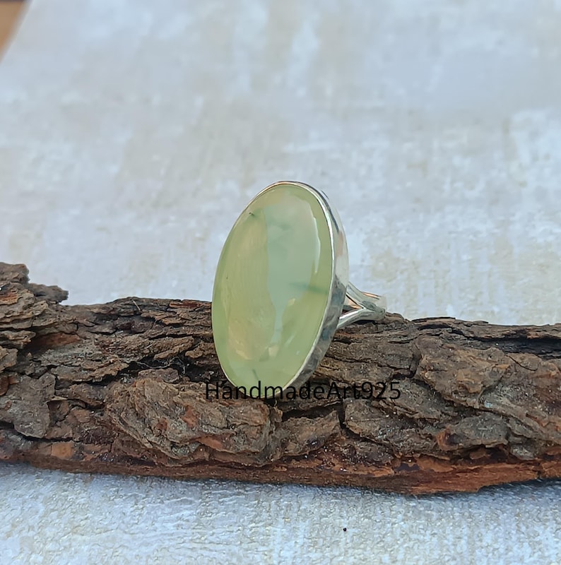 K&ouml;nnte beinhalten: Ein silberner Ring mit einem gro&szlig;en ovalen gr&uuml;nen Edelstein. Der Ring liegt auf einem St&uuml;ck Holz.