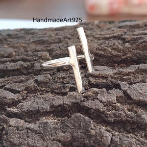 Op de afbeelding: Een zilveren ring met een modern design. De ring heeft twee verticale staven en een horizontale staaf, wat een unieke en minimalistische uitstraling creëert. De ring staat tegen een getextureerde, donkerbruine achtergrond. De tekst "HandmadeArt925" is zichtbaar.