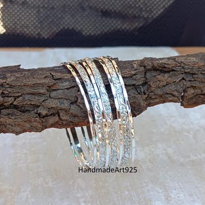 May include: Set of six silver bangles with a floral design. The bangles are stacked on a wooden surface.