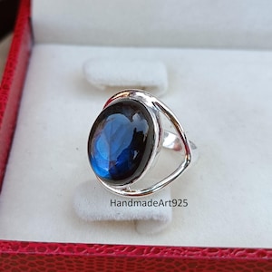 May include: A silver ring featuring a large, oval, blue gemstone. The gemstone has a dark, reflective surface. The ring is presented in a red jewellery box with a white interior. The text "HandmadeArt925" is visible.