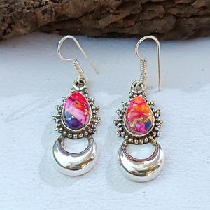 May include: A pair of silver dangle earrings. Each earring features a teardrop-shaped, multi-colored stone with red, orange, and purple hues, surrounded by a decorative silver setting. A crescent moon shape hangs below the stone.