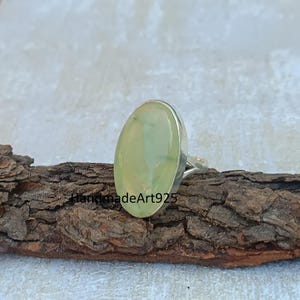 K&ouml;nnte beinhalten: Ein silberner Ring mit einem hellgr&uuml;nen ovalen Edelstein. Der Ring liegt auf einem St&uuml;ck Holz.