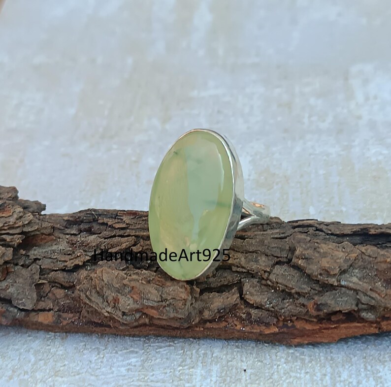K&ouml;nnte beinhalten: Ein silberner Ring mit einem hellgr&uuml;nen ovalen Edelstein. Der Ring liegt auf einem St&uuml;ck Holz.