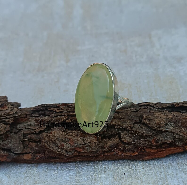 K&ouml;nnte beinhalten: Ein silberner Ring mit einem hellgr&uuml;nen ovalen Edelstein. Der Ring liegt auf einem St&uuml;ck Holz.