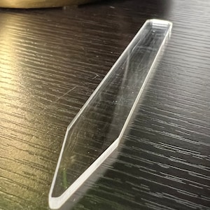 May include: A clear, long, and thin acrylic tool with a pointed tip. The tool is lying on a dark wood surface.