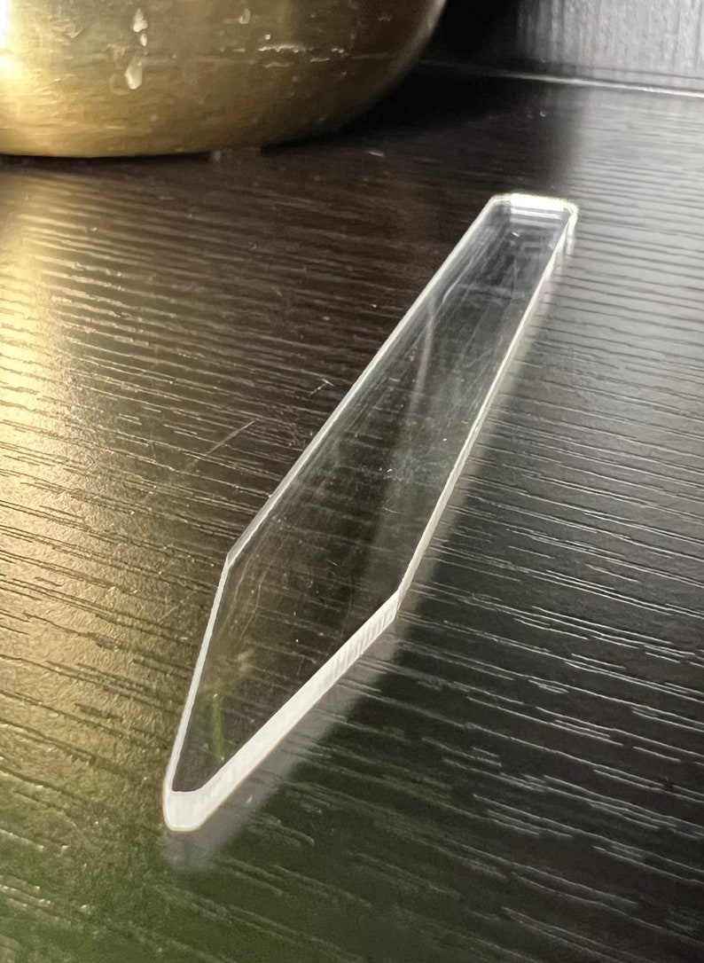 May include: A clear, long, and thin acrylic tool with a pointed tip. The tool is lying on a dark wood surface.