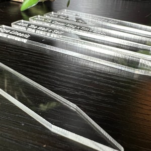 May include: Close-up of four clear acrylic plant markers with white text. The text reads "Anth. The Viall x Self", "Anth. T.A. Crystal Mag x Tim's Black", "Amth. Red Pip x Paul", and "Haya Linearis".