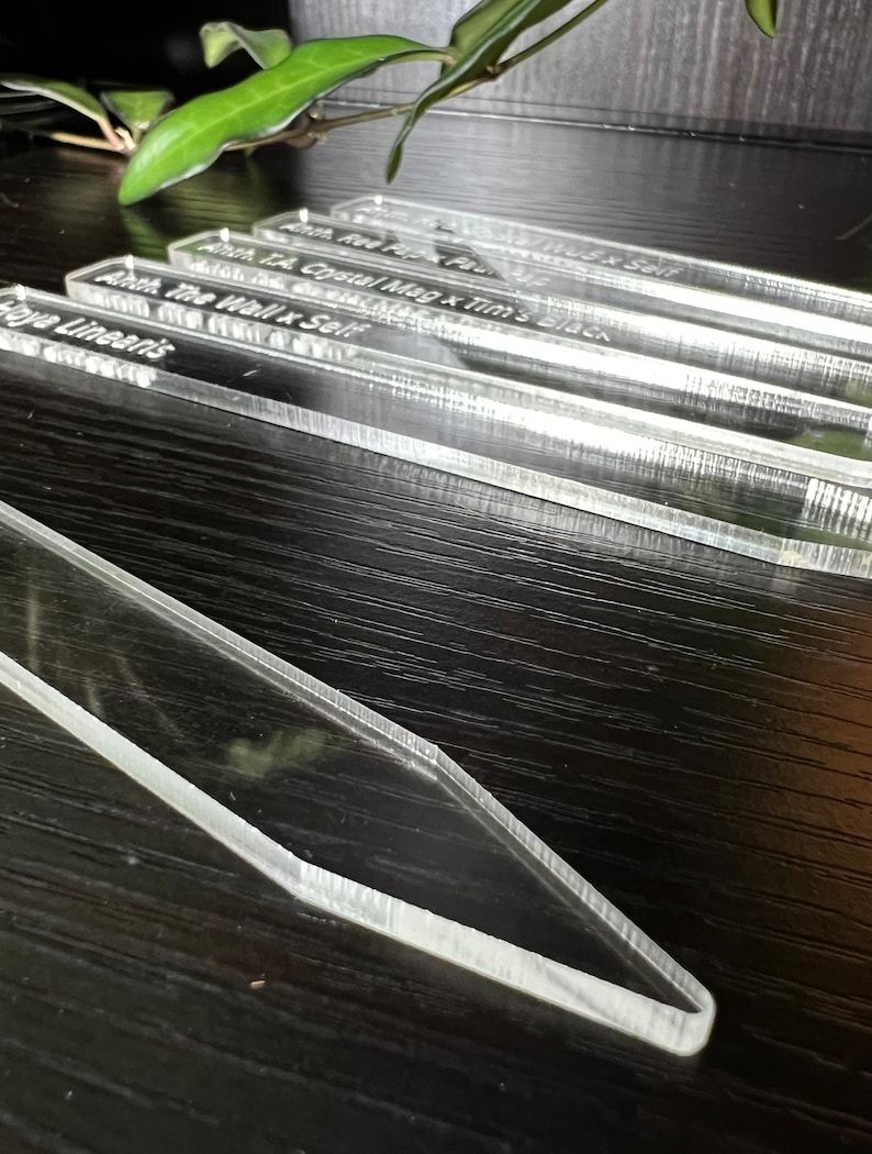 May include: Close-up of four clear acrylic plant markers with white text. The text reads "Anth. The Viall x Self", "Anth. T.A. Crystal Mag x Tim's Black", "Amth. Red Pip x Paul", and "Haya Linearis".