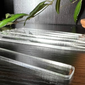 May include: A set of clear acrylic rulers on a black wooden surface. The rulers are stacked on top of each other and have a slight curve to them.