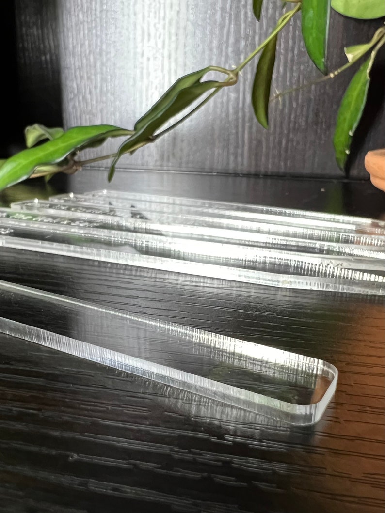 May include: A set of clear acrylic rulers on a black wooden surface. The rulers are stacked on top of each other and have a slight curve to them.