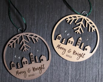 May include: Two wooden Christmas ornaments with a snowflake design and a cityscape silhouette. Both ornaments have the text "Merry & Bright 2022" on them.