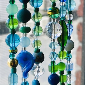 May include: Close-up of a beaded wind chime with a variety of blue, green, and clear glass beads. The beads are different shapes and sizes, including round, oval, and faceted. Some beads have gold accents. The wind chime is hanging in front of a window.