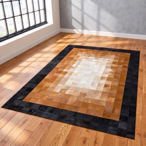 May include: A rectangular area rug constructed from a patchwork of individual square cowhide tiles. The rug features a central gradient pattern transitioning from white to light tan and brown, framed by a wide border of solid black tiles.
