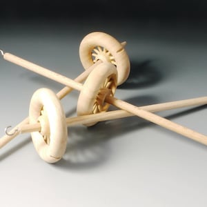 Könnte beinhalten: Drei Spinnräder aus Holz, jedes mit einem kreisförmigen Rad und einem langen, spitzen Schaft. Die Räder haben eine helle Holzoberfläche und ein dekoratives Speichendesign. Die Spindeln sind in verschiedenen Winkeln vor einem neutralen Hintergrund angeordnet.