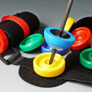 Pode incluir: Um pião de madeira azul com um centro preto e um eixo de metal. O pião é rodeado por discos de plástico coloridos em vermelho, verde, amarelo e azul. Os discos são presos a uma bolsa de tecido preto com uma alça de velcro.