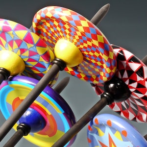 May include: Five colourful wooden spinning tops with geometric patterns on black sticks. The tops are red, yellow, blue, white and black.