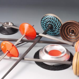 Op de afbeelding: Een verzameling handgemaakte spintollen in verschillende kleuren en ontwerpen. De spintollen hebben houten vliegwielen met geschilderde patronen, waaronder spiralen en strepen, en zijn gemonteerd op slanke assen. Sommige hebben feloranje of zilveren accenten.